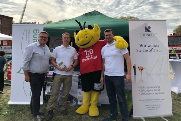 IMG_3646 Personen und ein Maskottchen an einem Veranstaltungsstand mit Stiftungsbannern.