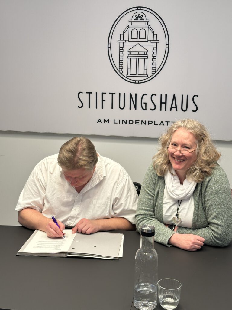 Eine Person unterzeichnet Unterlagen am Tisch, die daneben sitzende Person lächelt in die Kamera.