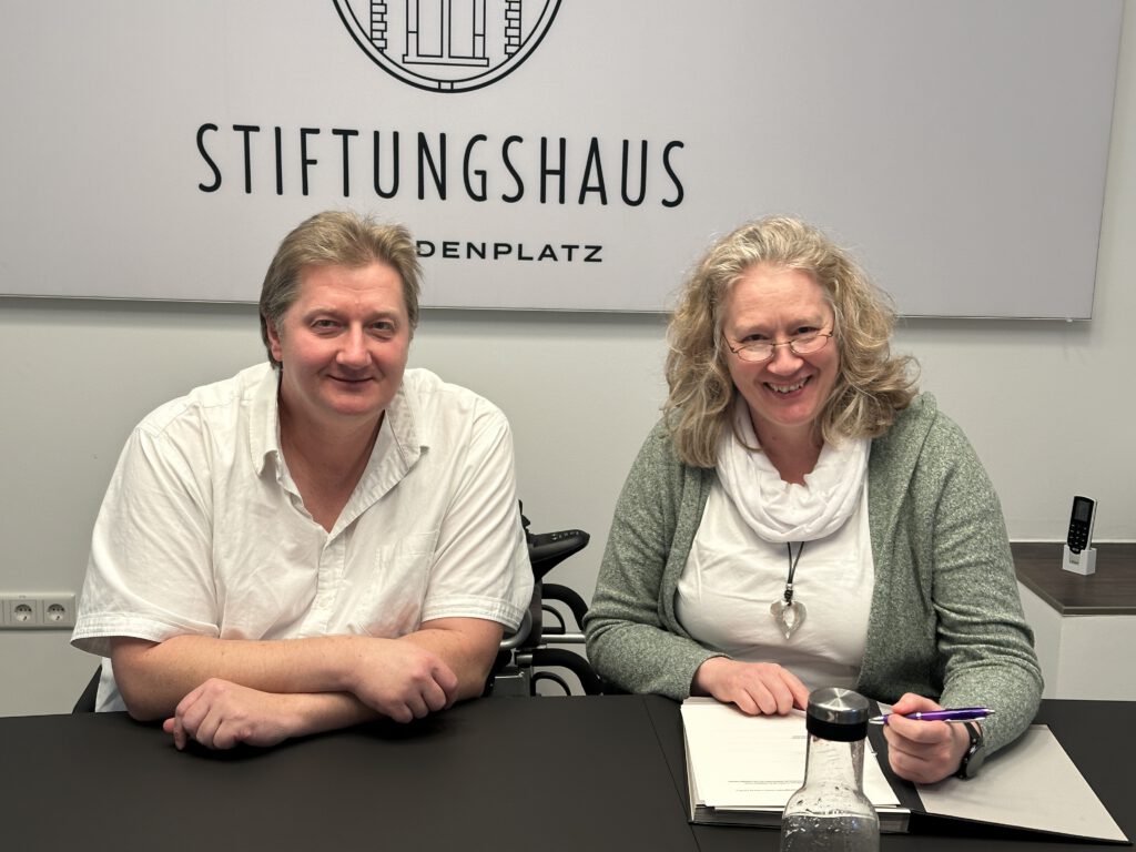 Zwei Personen sitzen nebeneinander am Tisch im Stiftungshaus am Lindenplatz und lächeln in die Kamera.